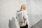 Frau mit einem hellgrauen urbanen Rolltop-Rucksack auf dem Rücken, seitliche Perspektive, vor einer Betonwand