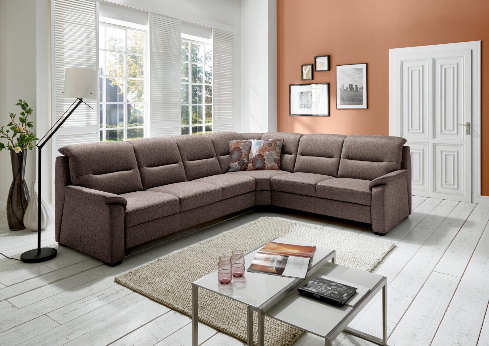 Große, moderne Polsterrundecke in einem hellen Wohnzimmer mit großen Fenstern, die viel Tageslicht hereinlassen. Die Couch ist in einem warmen Braunton gehalten und bietet viel Platz zum Entspannen. Perspektive von vorne links.