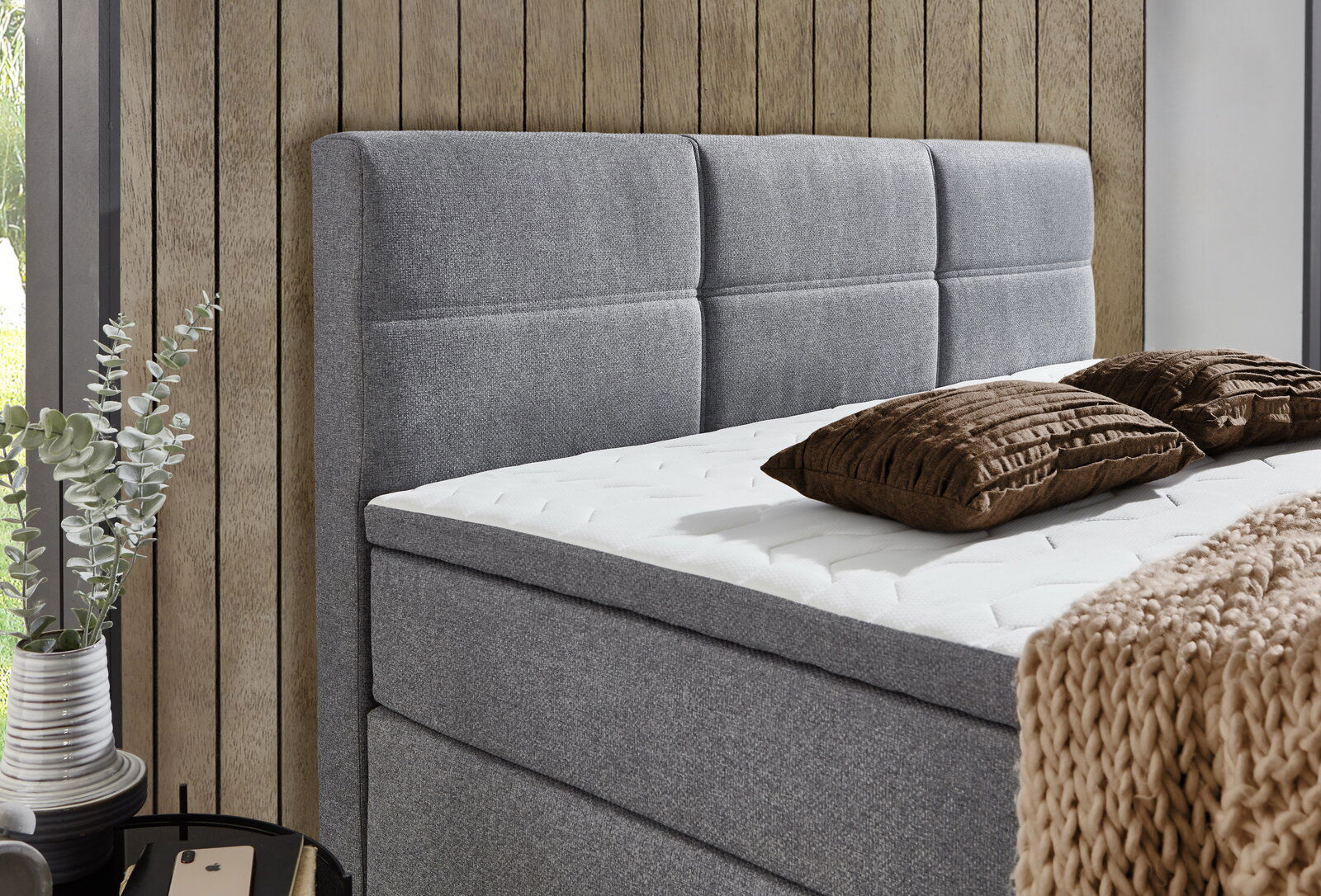 Seitliche Ansicht eines modernen grauen Boxspringbetts mit gepolstertem Kopfteil, dekoriert mit braunen Kissen und einer beigen Strickdecke.