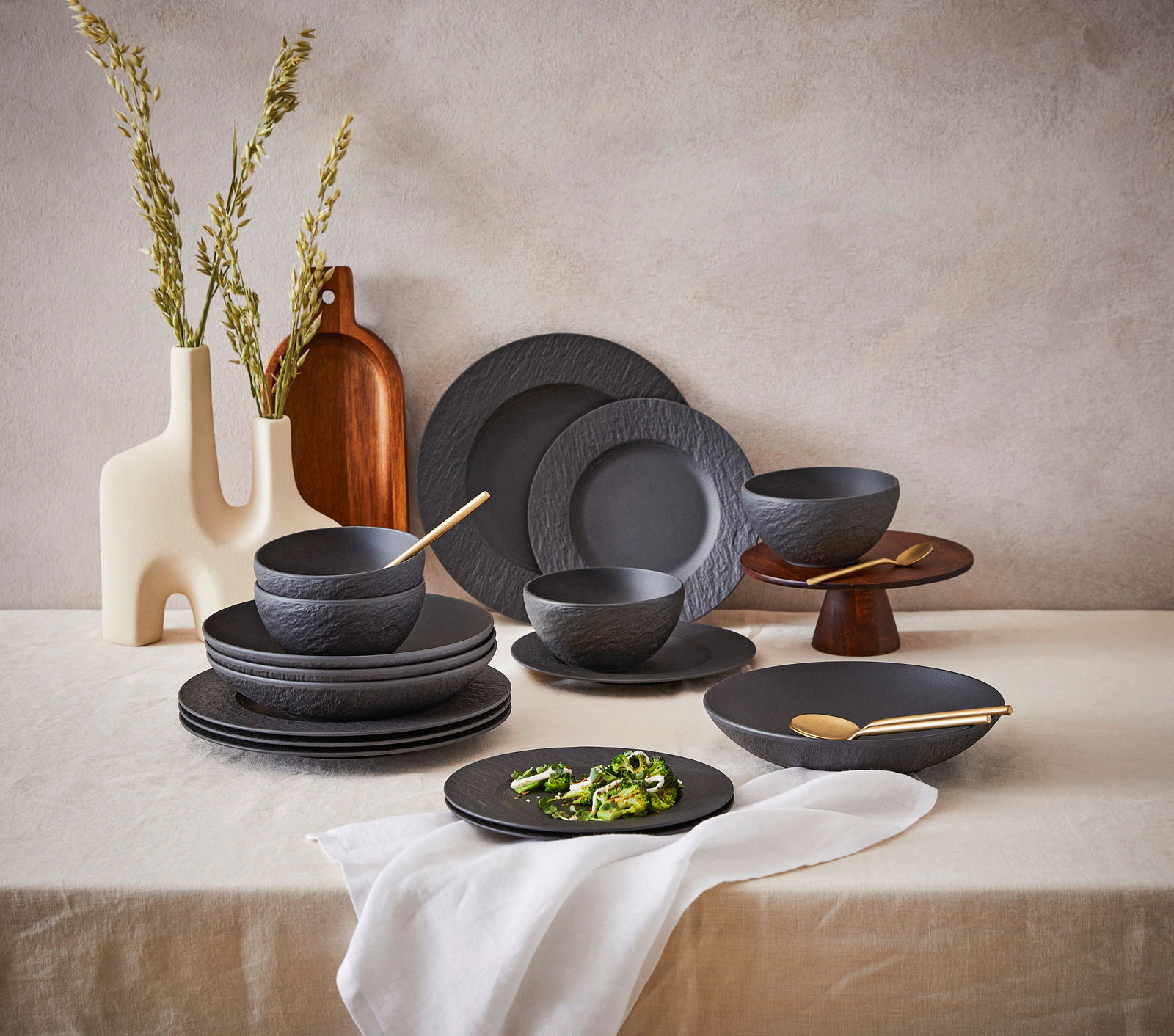 Villeroy & Boch Kombiservice 16-tlg. MANUFACTURE ROCK Elegantes 16-teiliges Geschirrset in mattem Schwarz mit natürlicher Steinoptik, bestehend aus Tellern und Schalen, auf einem Tisch mit dekorativen Vasen und Holzbrett, seitliche Perspektive.