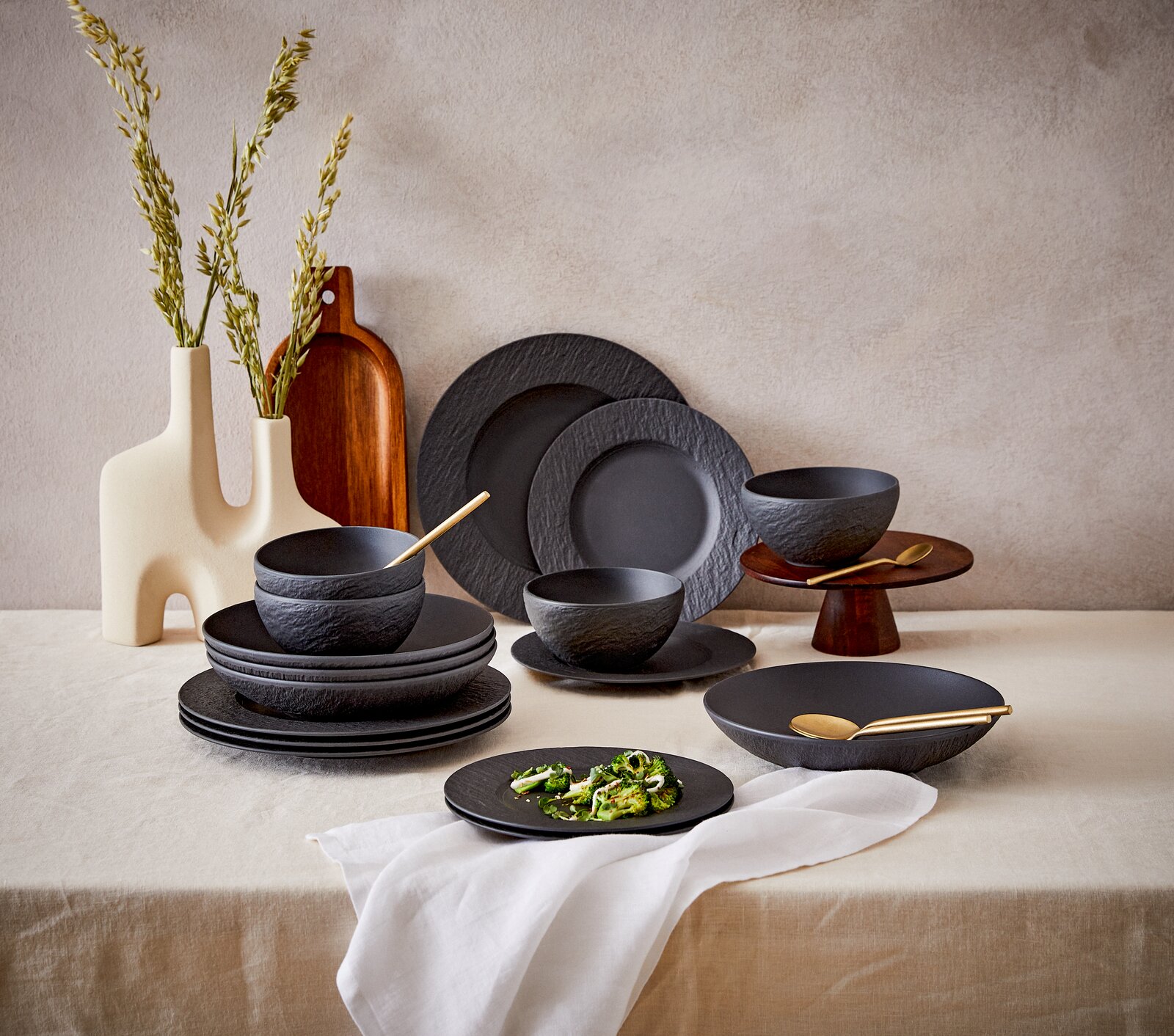 Villeroy & Boch Kombiservice 16-tlg. MANUFACTURE ROCK Elegantes 16-teiliges Geschirrset in mattem Schwarz mit natürlicher Steinoptik, bestehend aus Tellern und Schalen, auf einem Tisch mit dekorativen Vasen und Holzbrett, seitliche Perspektive.