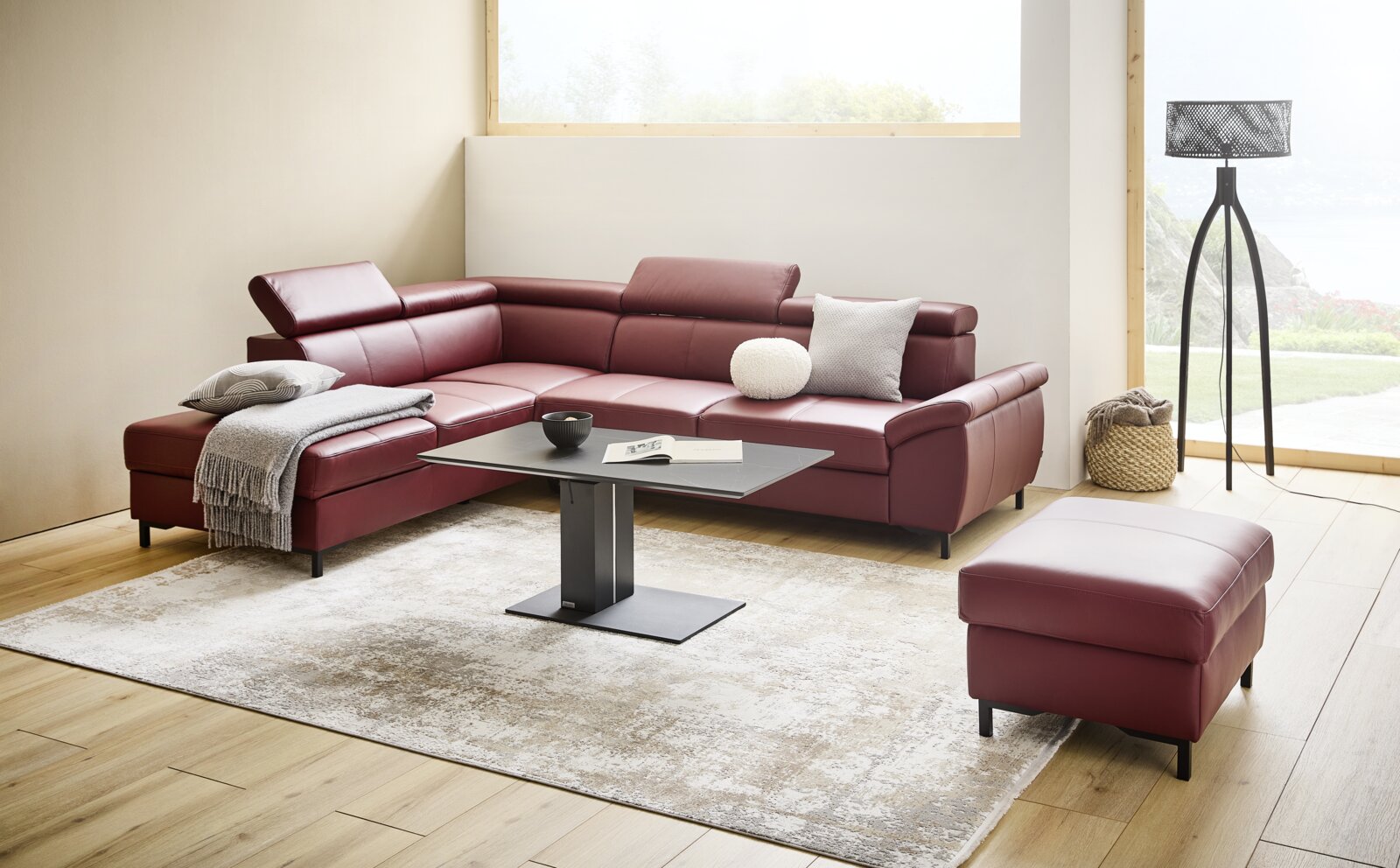 Stylife Ecksofa LENO 2.0 Moderne Eckgarnitur in weinrotem Leder mit verstellbaren Kopfstützen, dekoriert mit Kissen und Decke, in einem hellen Wohnzimmer mit großem Fenster und stilvoller Stehlampe, aus frontaler Perspektive.