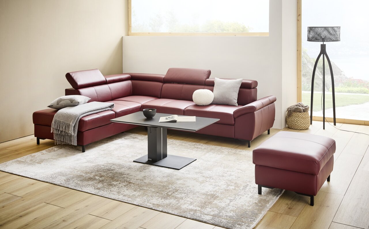 Stylife Ecksofa LENO 2.0 Moderne Eckgarnitur in weinrotem Leder mit verstellbaren Kopfstützen, dekoriert mit Kissen und Decke, in einem hellen Wohnzimmer mit großem Fenster und stilvoller Stehlampe, aus frontaler Perspektive.