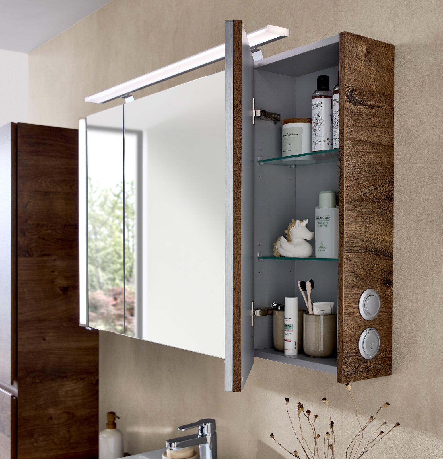 Geöffneter Badezimmerspiegelschrank mit Holzoptik-Seitenteil, integrierter LED-Aufsatzleuchte und Glasablagen mit Pflegeprodukten, aufgenommen aus schräger Frontansicht (leicht von rechts).