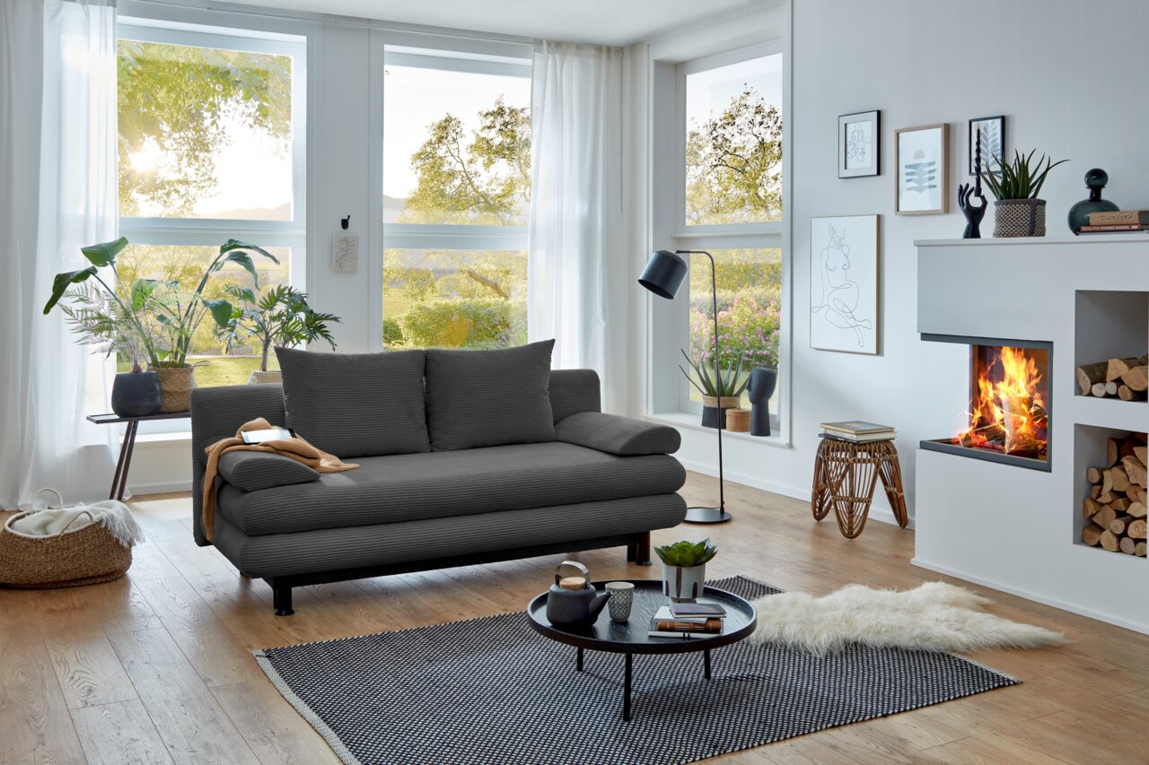 Graues Funktionssofa in modernem Wohnzimmer mit großen Fenstern, Pflanzen und Kamin, seitliche Perspektive.