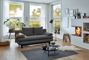 Graues Funktionssofa in modernem Wohnzimmer mit großen Fenstern, Pflanzen und Kamin, seitliche Perspektive.