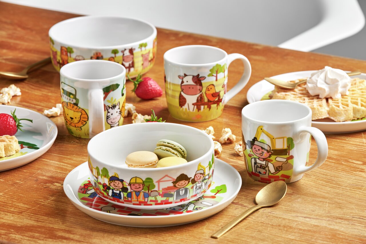 Kindergeschirr-Set mit Baustellenmotiven, bestehend aus Tasse, Schüssel und Teller, auf einem Holztisch arrangiert. Frontalansicht.