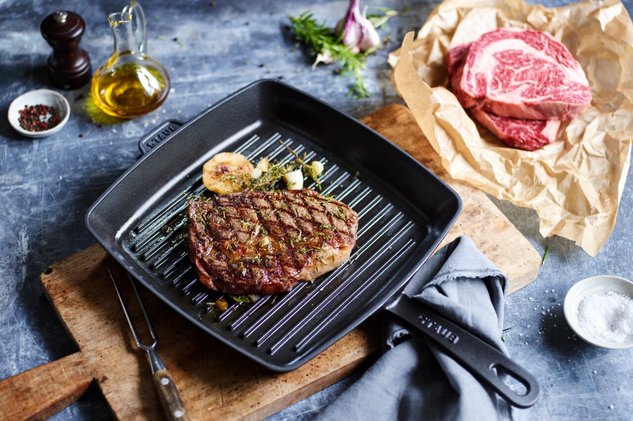 Grillpan van 26 x 26 cm met gegrilde biefstuk, knoflook en kruiden, gefotografeerd van bovenaf.