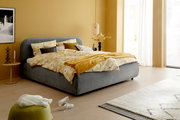 Graues Polsterbett in einem modern eingerichteten Schlafzimmer mit gelber Bettwäsche und Dekoration, seitliche Perspektive
