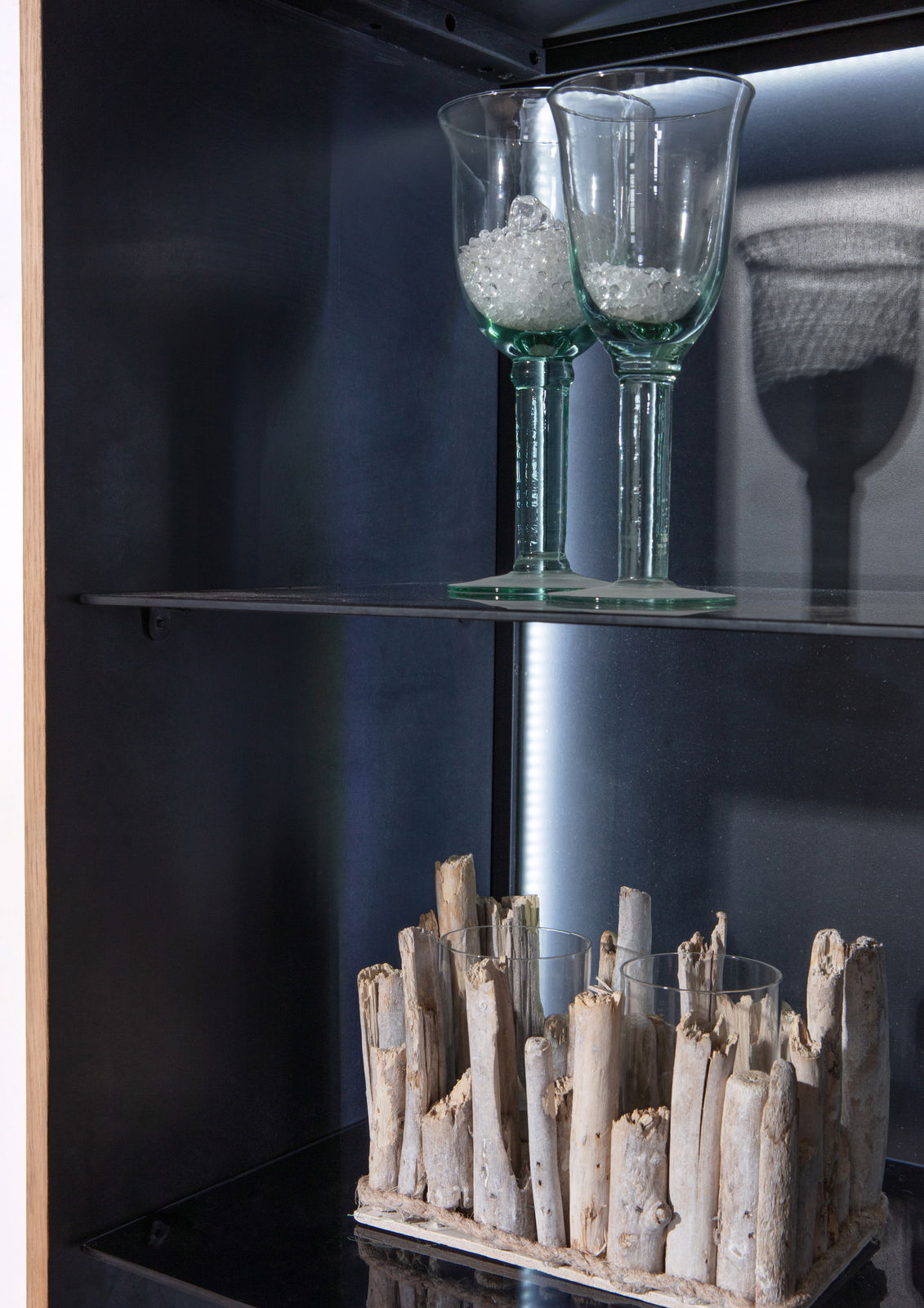 Nahaufnahme einer Vitrine mit zwei Gläsern auf einem Glasregal und einer dekorativen Holzstruktur darunter, seitliche Perspektive.