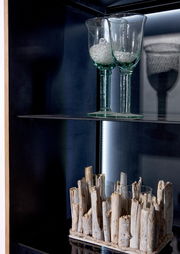 Nahaufnahme einer Vitrine mit zwei Gläsern auf einem Glasregal und einer dekorativen Holzstruktur darunter, seitliche Perspektive.