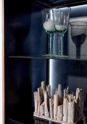 Nahaufnahme einer Vitrine mit zwei Gläsern auf einem Glasregal und einer dekorativen Holzstruktur darunter, seitliche Perspektive.