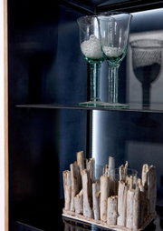 Nahaufnahme einer Vitrine mit zwei Gläsern auf einem Glasregal und einer dekorativen Holzstruktur darunter, seitliche Perspektive.