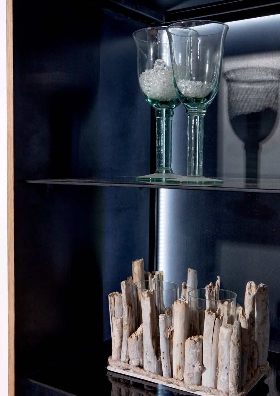 Close-up van een vitrinekast met twee glazen op een glazen plank en een decoratieve houten structuur eronder, gezien vanuit de zijkant.
