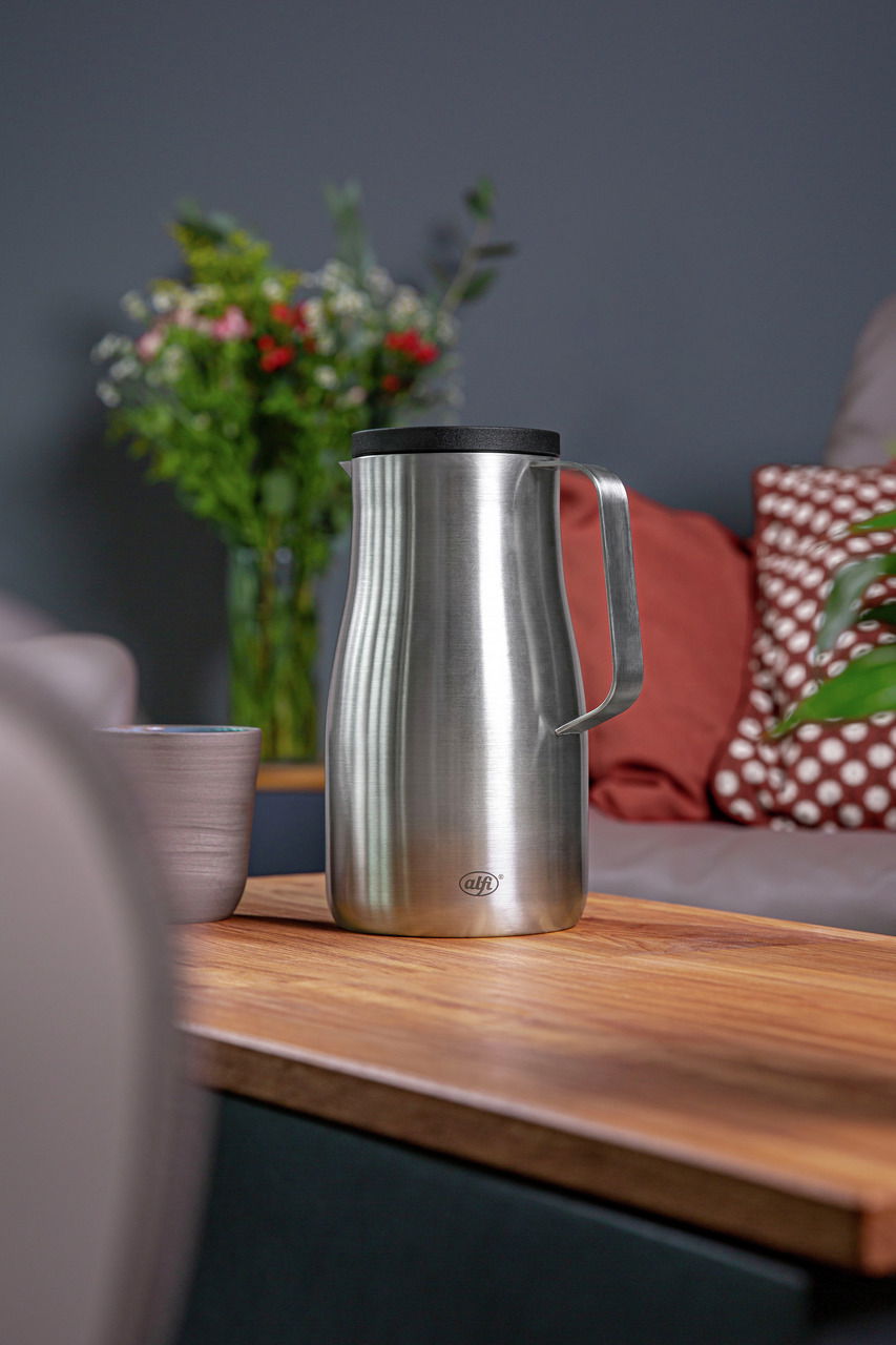 Isolierkanne Studio aus Edelstahl mit schwarzem Deckel, seitliche Perspektive auf einem Holztisch in einem gemütlichen Wohnzimmer.