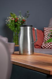 Isolierkanne Studio aus Edelstahl mit schwarzem Deckel, seitliche Perspektive auf einem Holztisch in einem gemütlichen Wohnzimmer.