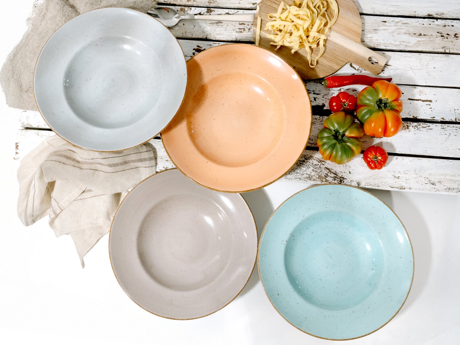 Draufsicht auf vier gesprenkelte Keramik-Speiseteller in Pastellfarben (Grau, Apricot, Beige, Hellblau) mit braunem Rand auf rustikalem Holztisch, dekoriert mit Schneidebrett, Nudeln und Tomaten.