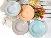 Draufsicht auf vier gesprenkelte Keramik-Speiseteller in Pastellfarben (Grau, Apricot, Beige, Hellblau) mit braunem Rand auf rustikalem Holztisch, dekoriert mit Schneidebrett, Nudeln und Tomaten.