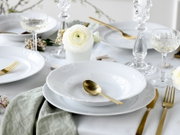 Schlicht gedeckter Esstisch mit weißem Porzellan-Geschirr (Teller und tiefer Teller), goldfarbenem Besteck, Kristallgläsern und Kerzenhaltern sowie weißer Blume, Perspektive: leicht von oben in schräger Seitenansicht.