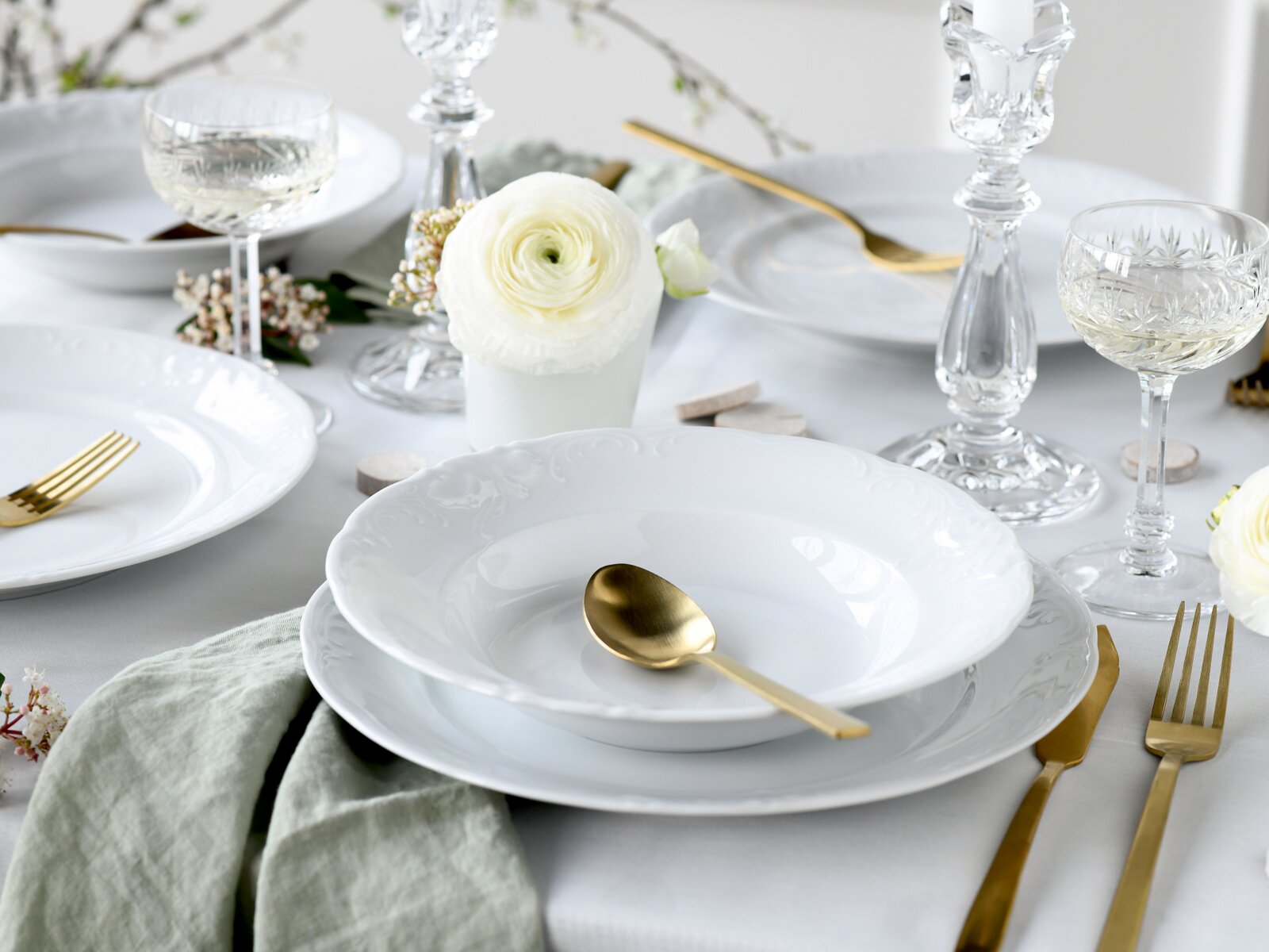 Schlicht gedeckter Esstisch mit weißem Porzellan-Geschirr (Teller und tiefer Teller), goldfarbenem Besteck, Kristallgläsern und Kerzenhaltern sowie weißer Blume, Perspektive: leicht von oben in schräger Seitenansicht.