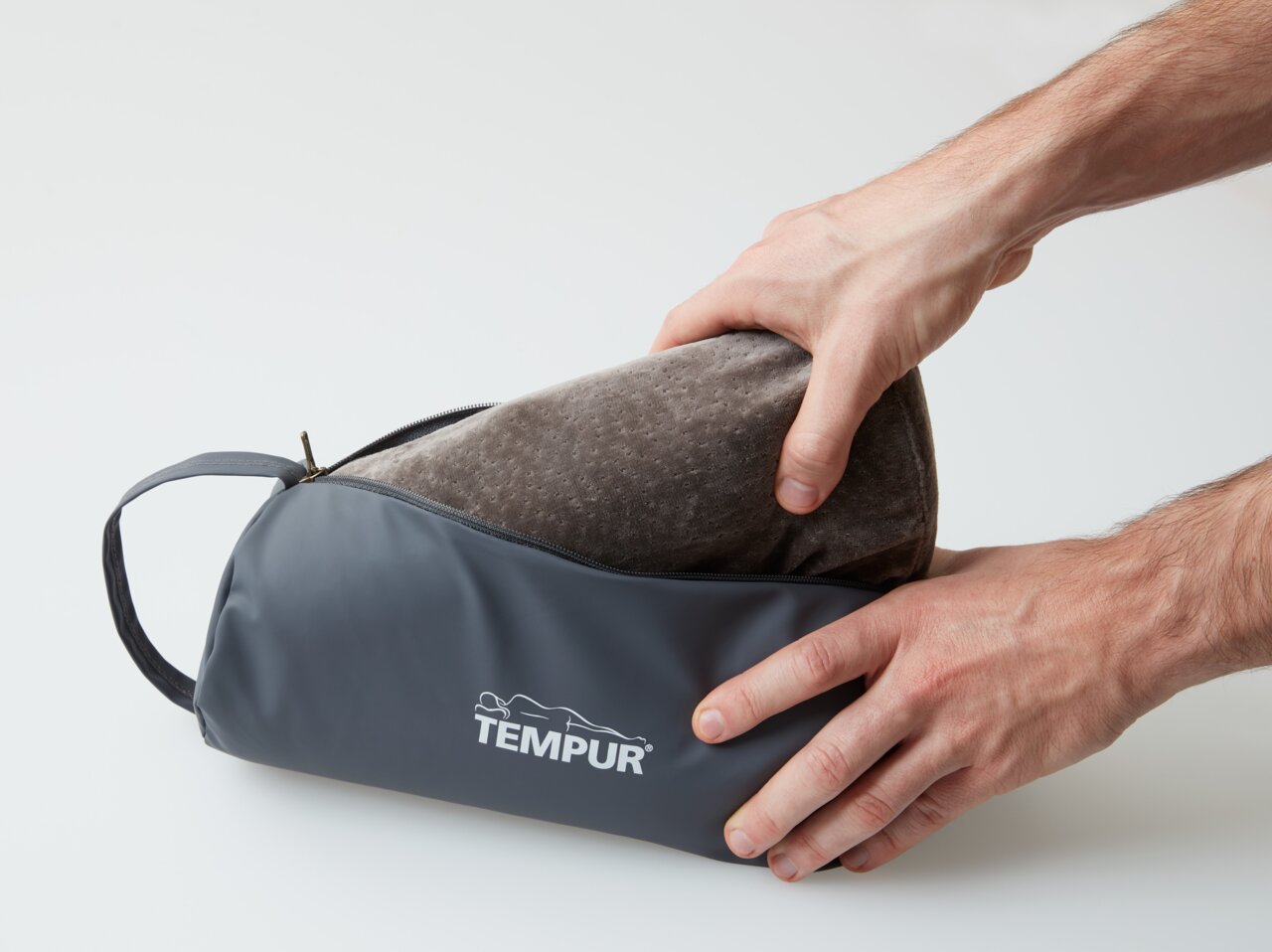 Tempur-reiskussen in een grijze tas, vastgehouden door twee handen, zijaanzicht