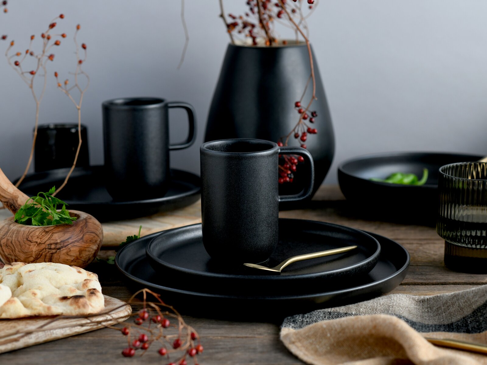 Mattschwarze Keramiktasse mit Henkel auf schwarzen Tellern, dekoratives Geschirr-Setting mit Vase und Beerenzweigen auf Holztisch, Perspektive: frontale Nahaufnahme auf Augenhöhe