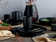 Mattschwarze Keramiktasse mit Henkel auf schwarzen Tellern, dekoratives Geschirr-Setting mit Vase und Beerenzweigen auf Holztisch, Perspektive: frontale Nahaufnahme auf Augenhöhe
