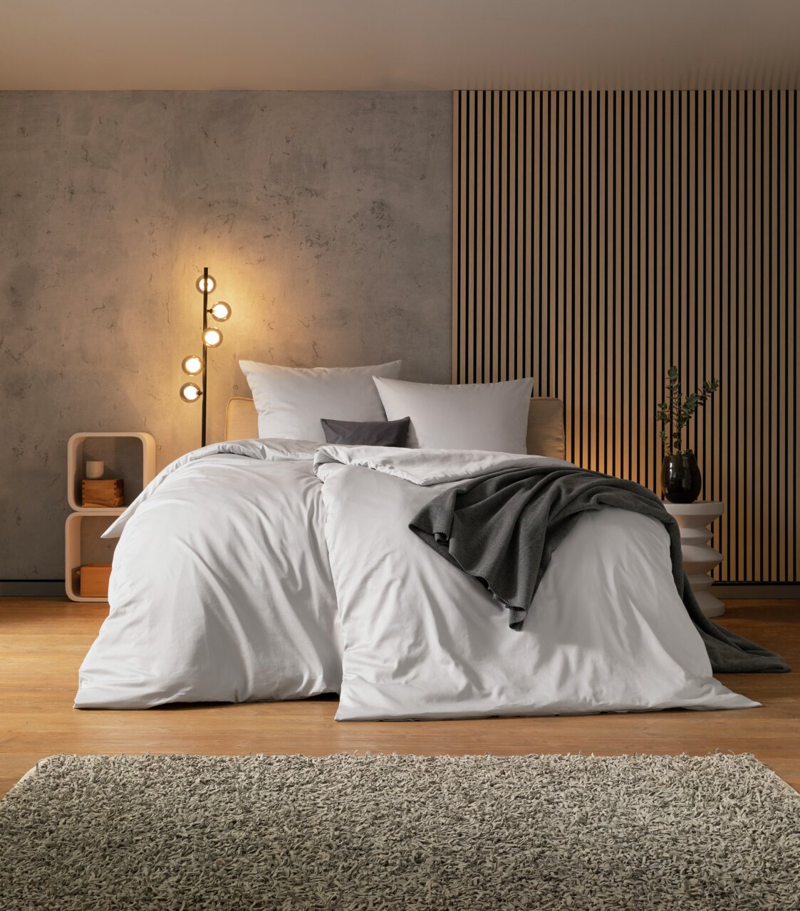 Modernes Schlafzimmer mit einem stilvollen Doppelbett, das mit weißen Bettbezügen und einem grauen Überwurf dekoriert ist. Im Hintergrund eine Betonwand und eine Lampe mit warmem Licht. Perspektive von vorne.