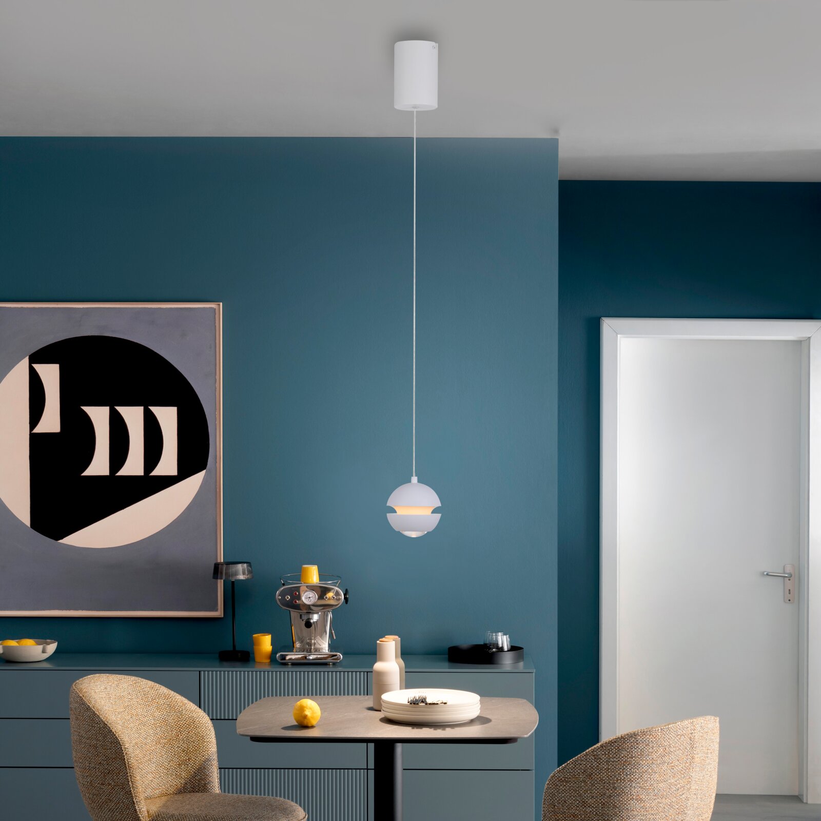 Moderne weiße Pendelleuchte mit kugelförmigem Lampenschirm und langem Kabel über einem Esstisch vor petrolfarbener Wand, Frontansicht auf Augenhöhe.