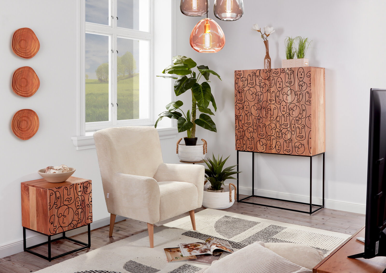 Modernes Wohnzimmer-Setting mit beigem Sessel, Holz-Sideboard und kleinem Beistellschrank mit grafischem Gesichter-Muster auf schwarzem Metallgestell, Pflanzen und Pendelleuchten; Perspektive: frontale, leicht seitliche Raumansicht.