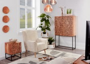 Moderne woonkamerinrichting met een beige fauteuil, een houten dressoir en een klein bijzetkastje met een grafisch gezichtspatroon op een zwart metalen onderstel, planten en hanglampen; perspectief: frontaal, licht zijdelings aanzicht van de ruimte.