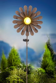 Solarleuchte in Form einer Blume mit gelbem Licht in der Mitte, auf einem Stab montiert, vor einem Hintergrund aus grünen Pflanzen, seitliche Perspektive.