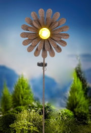 Zonne-lamp in de vorm van een bloem met geel licht in het midden, gemonteerd op een stok, tegen een achtergrond van groene planten, zijaanzicht.