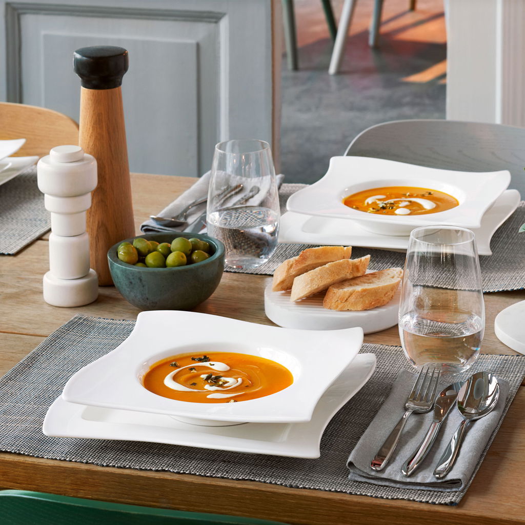 Eleganter Speiseteller 27cm NewWave mit geschwungenem Design, präsentiert auf einem gedeckten Tisch mit Suppe, Brot und Besteck, aus der Vogelperspektive.