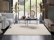 Moderner Wohnraum mit hellem Wollteppich, der vor einem Sofa und einem Couchtisch liegt. Der Raum ist stilvoll eingerichtet mit großen Fenstern im Hintergrund. Perspektive von vorne auf den Teppich.