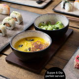 Zwei schwarze Schalen mit Suppe und Gemüse auf einem Holzbrett, umgeben von Sushi auf einem Tisch, aus der Vogelperspektive.