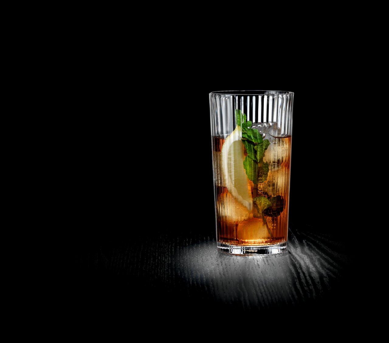 Ein großer, geriffelter Glasbecher mit einem erfrischenden Getränk, Eiswürfeln, Zitronenscheibe und Minzblättern, auf einer dunklen Oberfläche stehend, seitliche Perspektive.