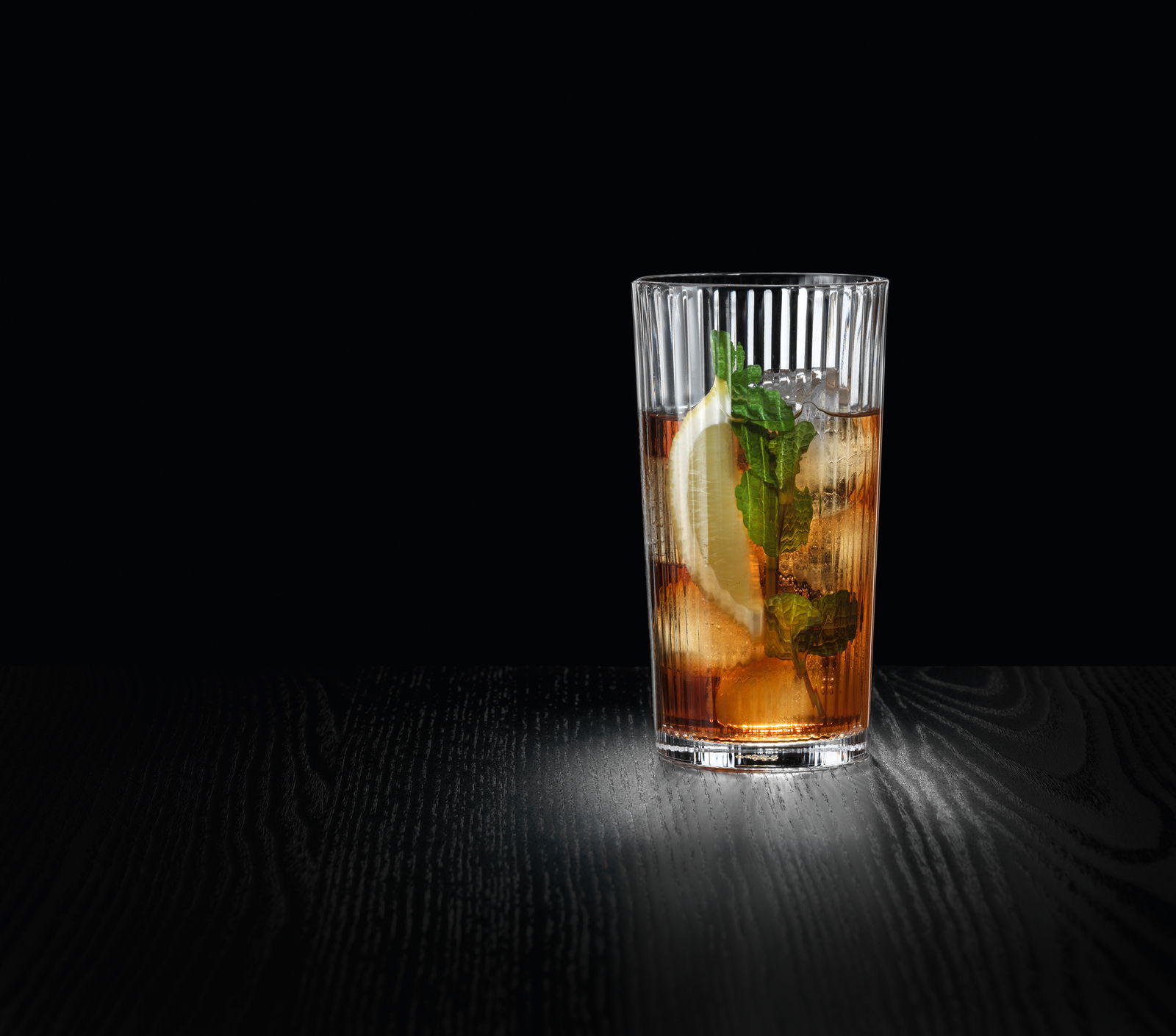 Ein großer, geriffelter Glasbecher mit einem erfrischenden Getränk, Eiswürfeln, Zitronenscheibe und Minzblättern, auf einer dunklen Oberfläche stehend, seitliche Perspektive.