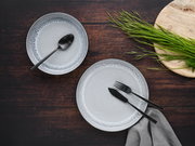 Zwei graue Teller mit schwarzem Besteck auf dunklem Holztisch, fotografiert aus der Vogelperspektive, mit grünem Gras auf einem Holzbrett.