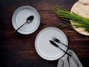 Zwei graue Teller mit schwarzem Besteck auf dunklem Holztisch, fotografiert aus der Vogelperspektive, mit grünem Gras auf einem Holzbrett.
