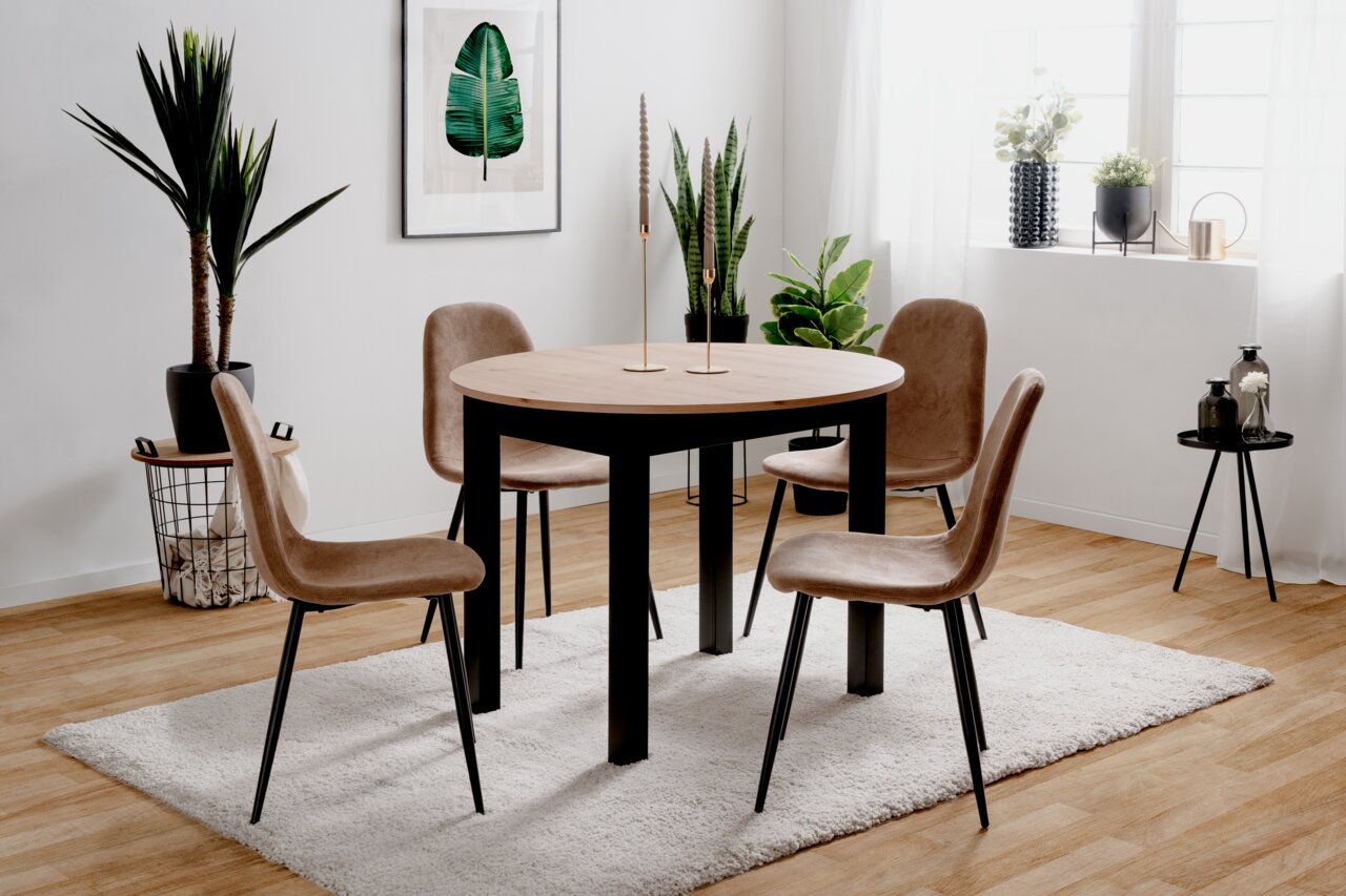 Z2 Funktions-Esstisch rund COBURG Moderner Esstisch mit runder Holzplatte und schwarzen Beinen, umgeben von vier beigen Stühlen, in einem hellen Raum mit Pflanzen und Dekorationen, seitliche Perspektive.