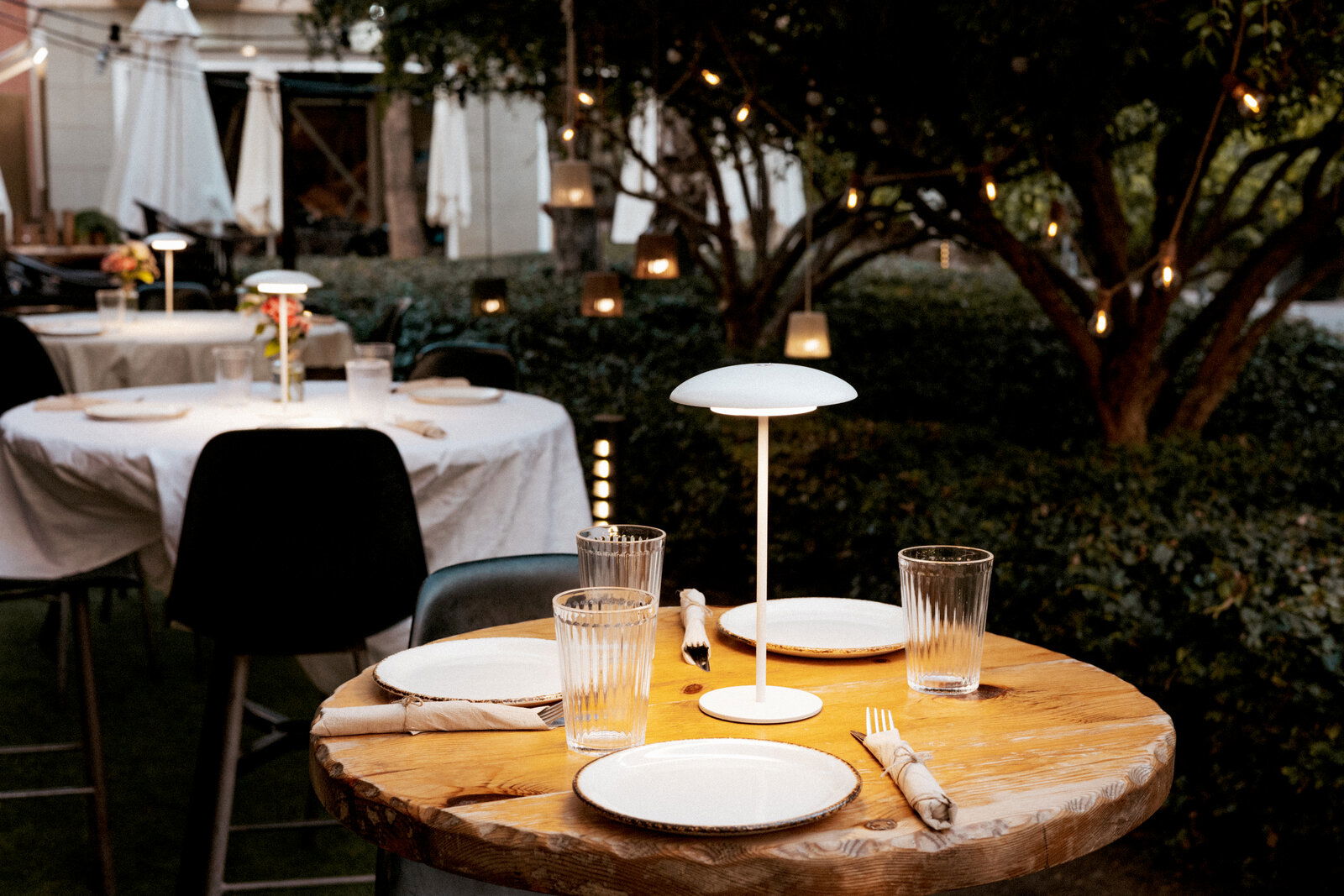 Schlanke, moderne Tischlampe mit rundem Schirm auf gedecktem Holztisch in einem stimmungsvoll beleuchteten Outdoor-Restaurantgarten, aufgenommen aus leicht erhöhter, frontaler Perspektive.