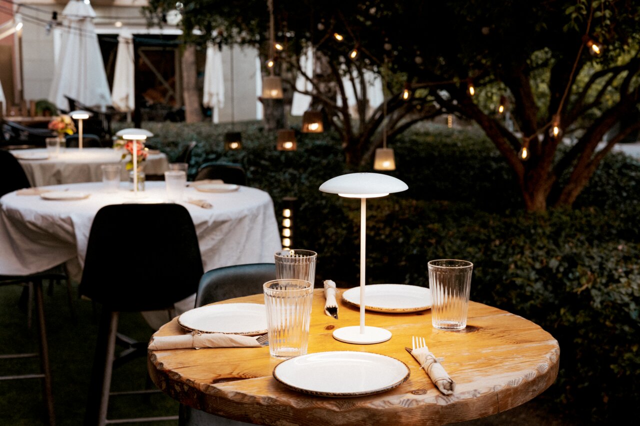 Schlanke, moderne Tischlampe mit rundem Schirm auf gedecktem Holztisch in einem stimmungsvoll beleuchteten Outdoor-Restaurantgarten, aufgenommen aus leicht erhöhter, frontaler Perspektive.