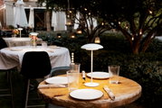 Schlanke, moderne Tischlampe mit rundem Schirm auf gedecktem Holztisch in einem stimmungsvoll beleuchteten Outdoor-Restaurantgarten, aufgenommen aus leicht erhöhter, frontaler Perspektive.