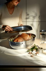 Eine Person hebt den Metalldeckel eines glänzenden Extrabräters an, in dem ein Braten auf einem Bett aus Kräutern liegt. Die Perspektive zeigt den Bräter von der Seite auf einer Küchenarbeitsplatte.