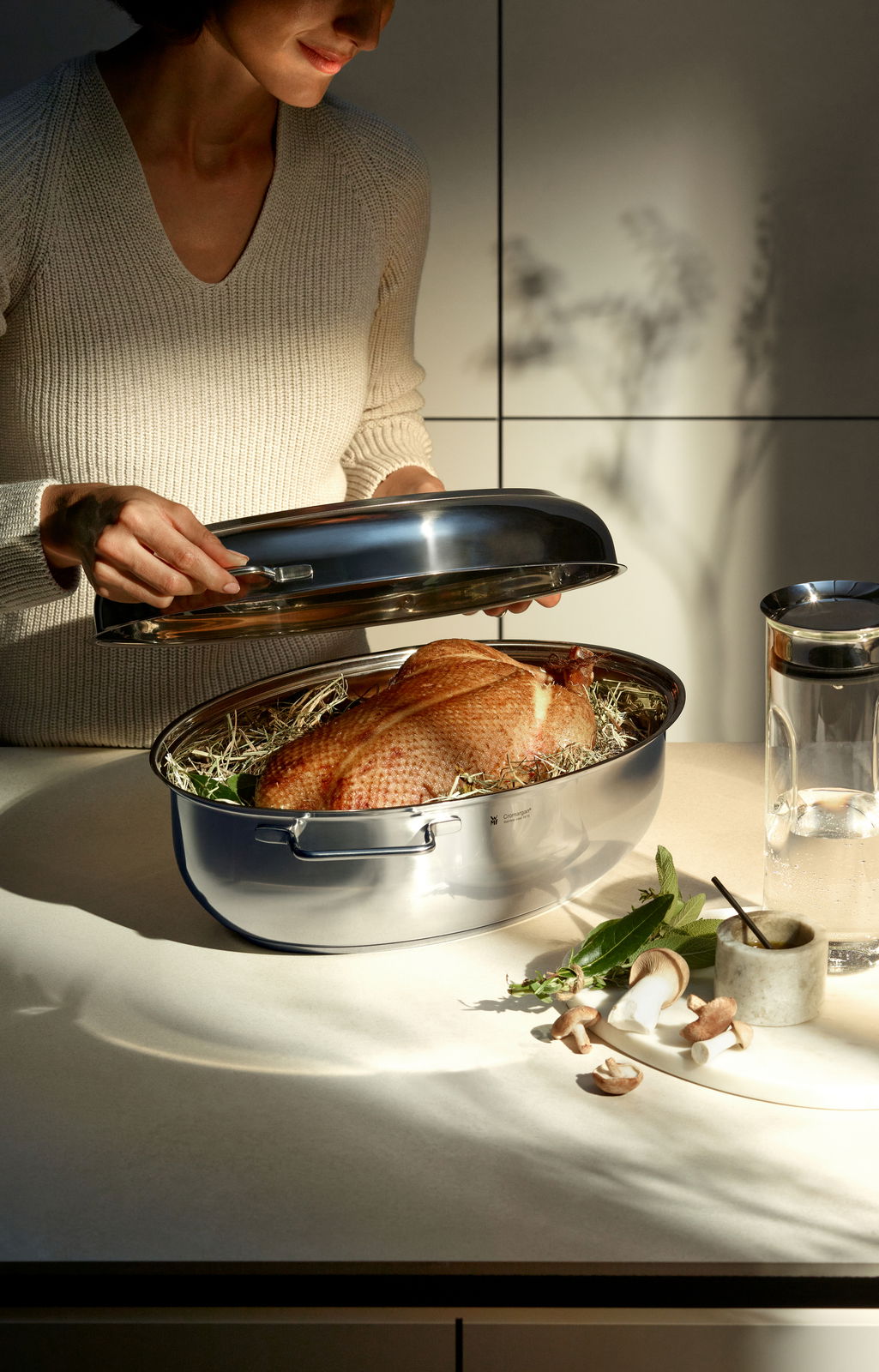 Eine Person hebt den Metalldeckel eines glänzenden Extrabräters an, in dem ein Braten auf einem Bett aus Kräutern liegt. Die Perspektive zeigt den Bräter von der Seite auf einer Küchenarbeitsplatte.