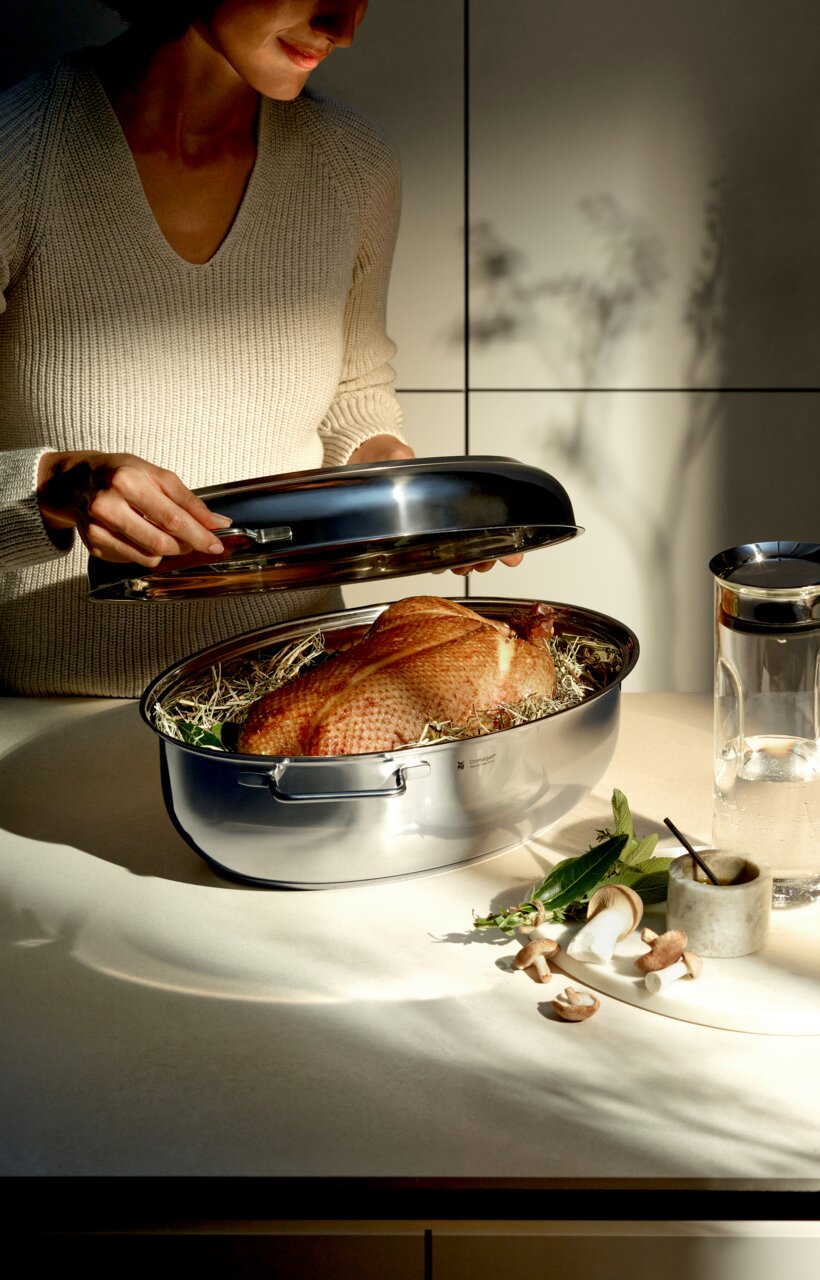 WMF Extrabräter Eine Person hebt den Metalldeckel eines glänzenden Extrabräters an, in dem ein Braten auf einem Bett aus Kräutern liegt. Die Perspektive zeigt den Bräter von der Seite auf einer Küchenarbeitsplatte.