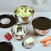 Roestvrijstalen mengkomset met zwarte deksels en rasp-inzet op een keukenblad, met salade, geraspte kaas en groenten eromheen, gefotografeerd in licht schuin bovenaanzicht.