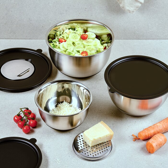 Roestvrijstalen mengkomset met zwarte deksels en rasp-inzet op een keukenblad, met salade, geraspte kaas en groenten eromheen, gefotografeerd in licht schuin bovenaanzicht.