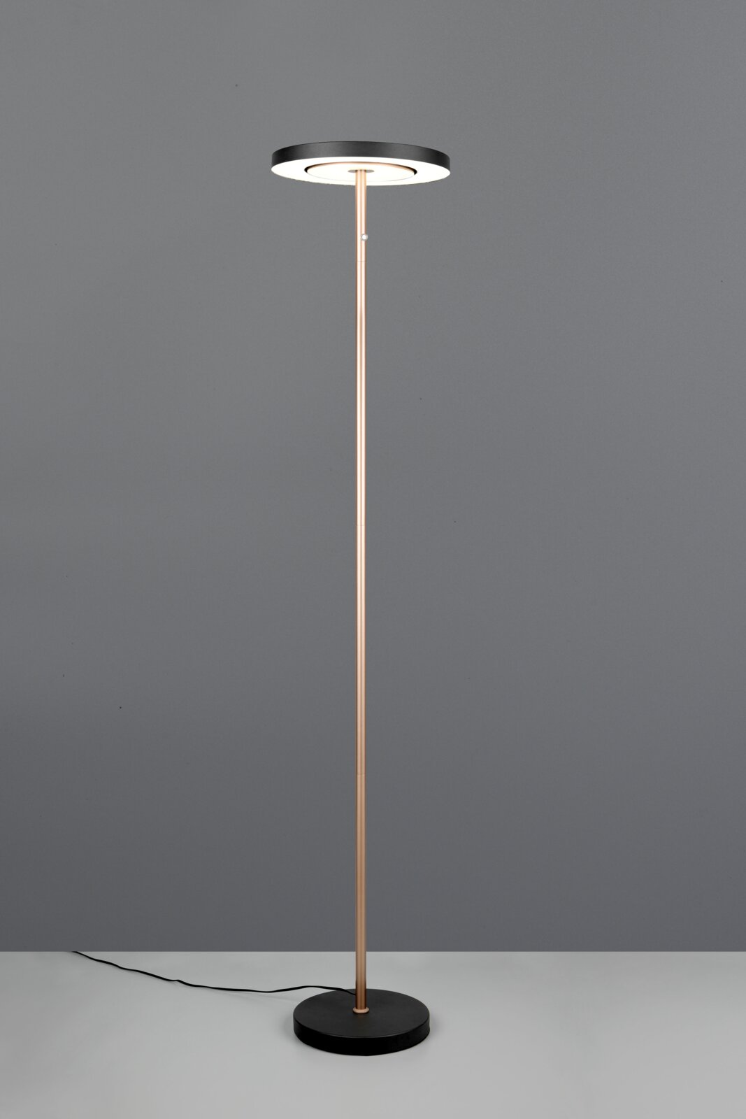 Moderne staande vloerlamp met slanke messingkleurige staander, ronde zwarte voet en ronde LED-lampkop, gefotografeerd van voren op ooghoogte tegen een grijze achtergrond